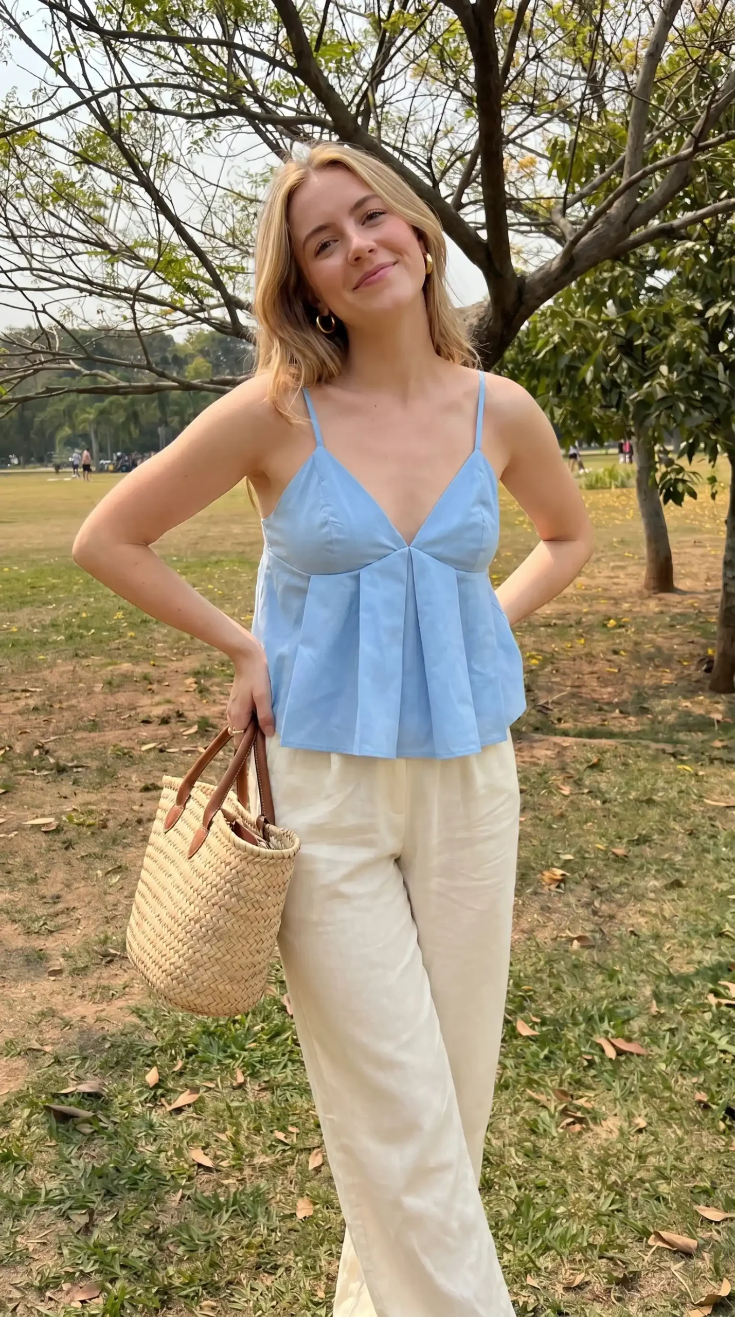 Woman wearing light blue pleated babydoll top with cream linen pants and woven tote outdoors
